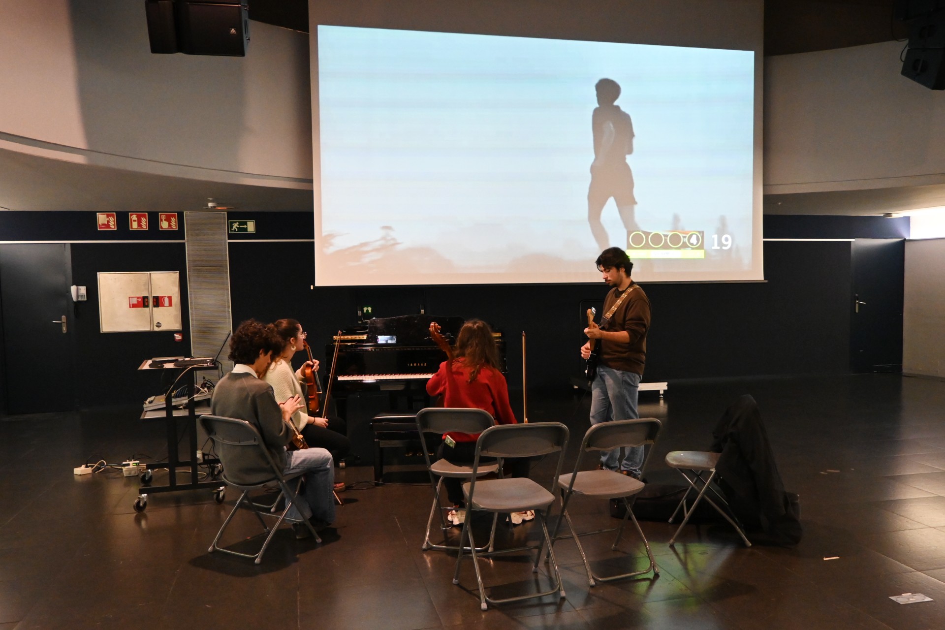Un dels grups toca instruments en una de les classes pràctiques a la Sala Aranyó del campus del Poblenou de la UPF. A la pantalla, un fotograma del metratge apropiat per al qual han de crear una banda sonora.
