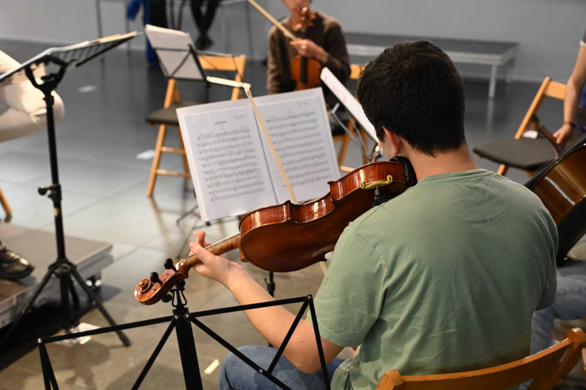 Un jove músic toca el violí durant un assaig de música de cambra. Està assegut d’esquena, concentrat en la partitura col·locada al faristol davant seu. Al fons, altres intèrprets també preparen els seus instruments en una sala d’assaig lluminosa.