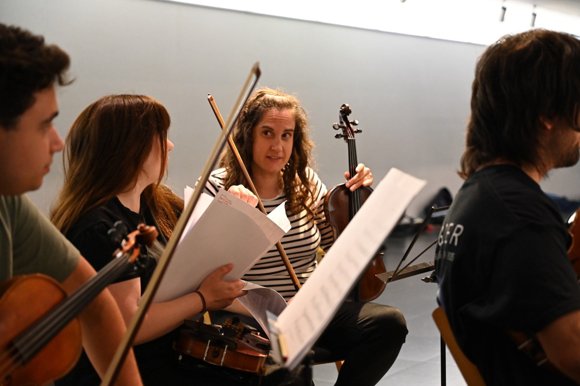 Quatre músics de corda asseguen en rotllana durant un assaig, intercanviant comentaris i partitures. Al centre, una dona amb una samarreta de ratlles sosté un violí i parla amb els altres membres, mentre els fulls de música cobreixen parcialment els instruments.